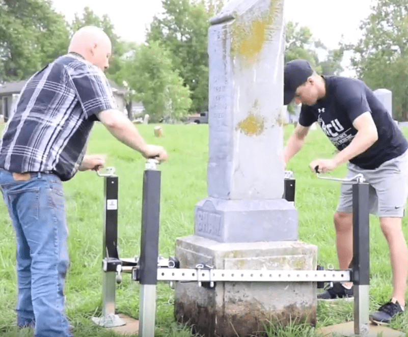 Two men are working to level a tilted gravestone using a Tombstone Jack and other mechanical tools in a grassy cemetery.